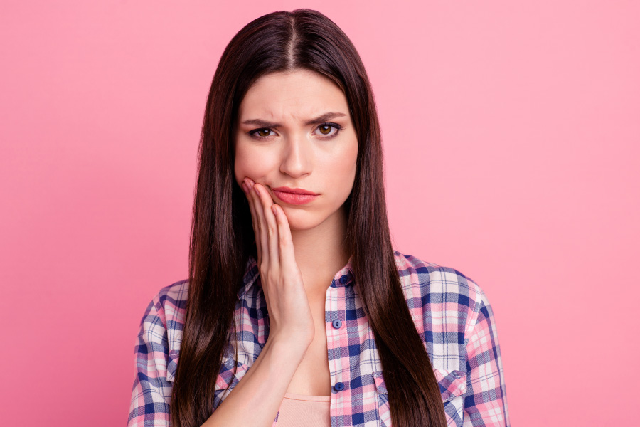 Brunette woman in a plaid shirt cringes in pain and touches her cheek because she needs root canal therapy