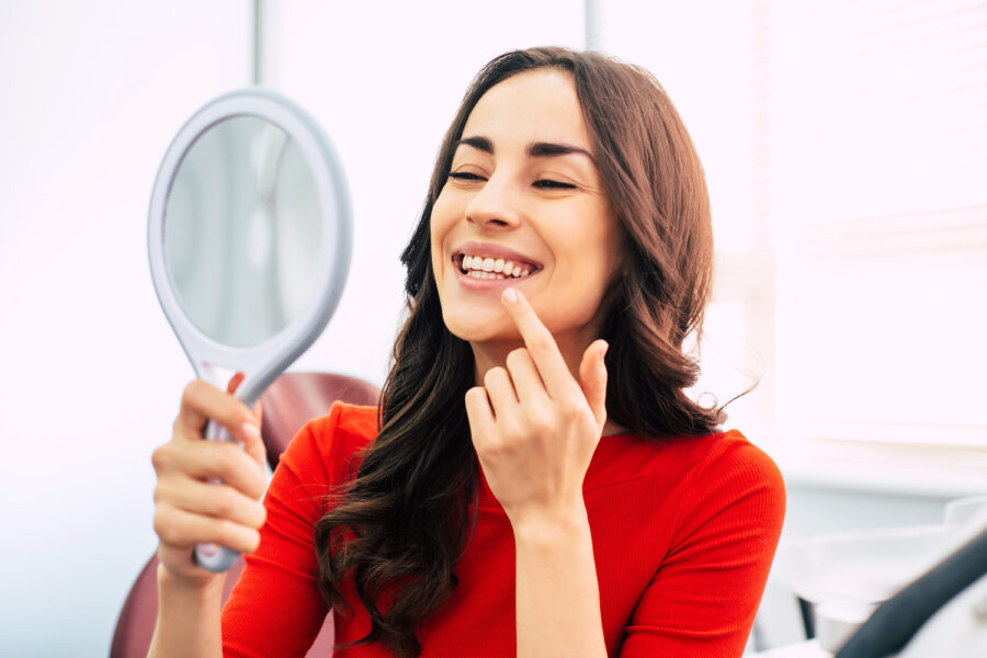 brunette woman in a red blouse smiles at herself in a handheld mirror while pointing to her teeth