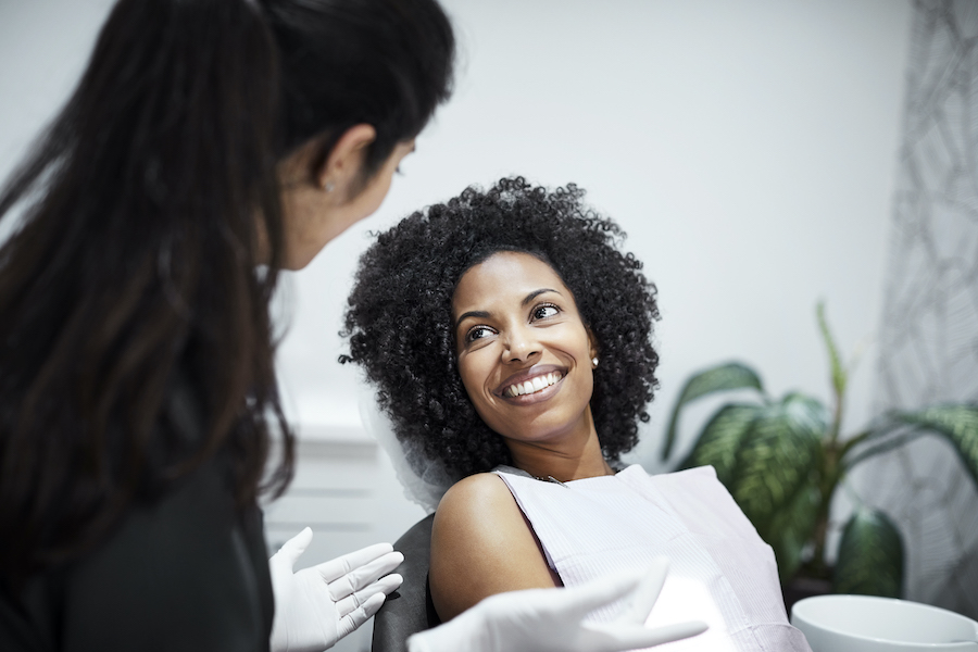 A curly-haired Black woman smiles with a white bib while sitting in a dental chair talking to her female dentist